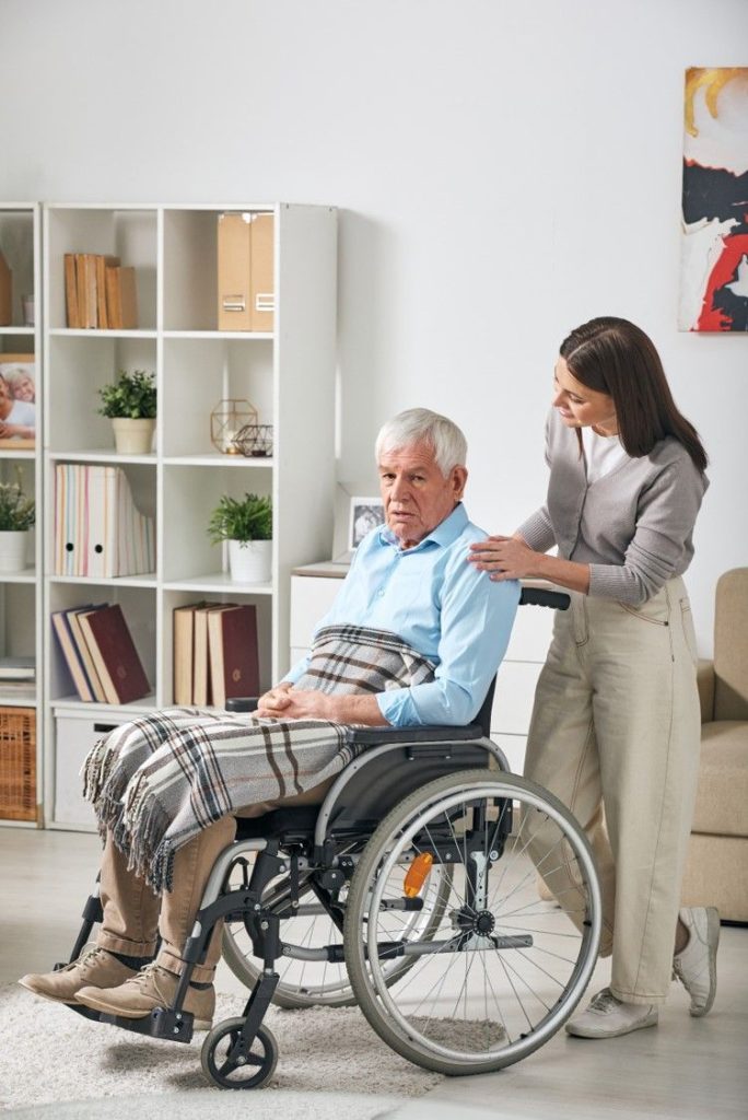 A carer with her hand on the shoulder of an older man in a wheelchair at home