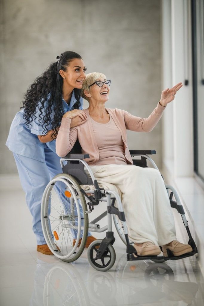 A carer and a smiling older woman in a wheelchair looking out of a window together
