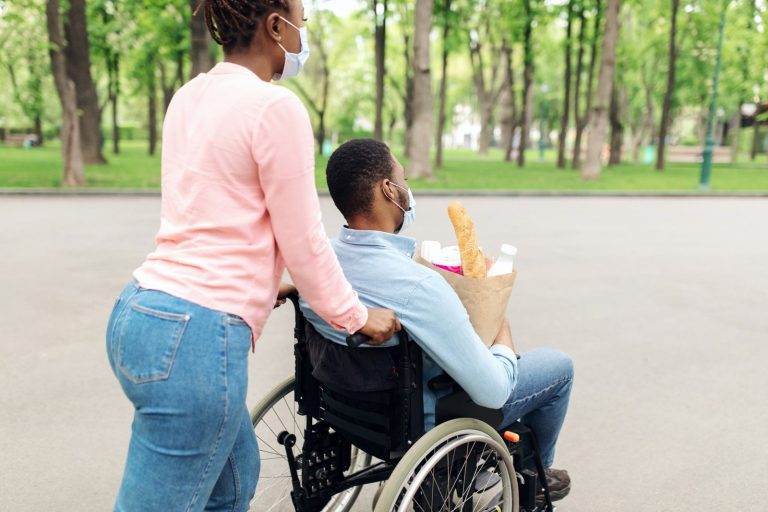 A carer pushing a man in a wheelchair through a park on the way back from the shops