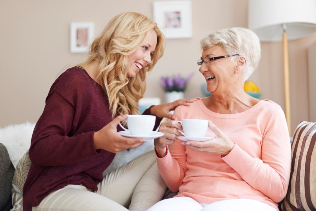 A carer and an older woman laughing together over cups of tea on the sofa